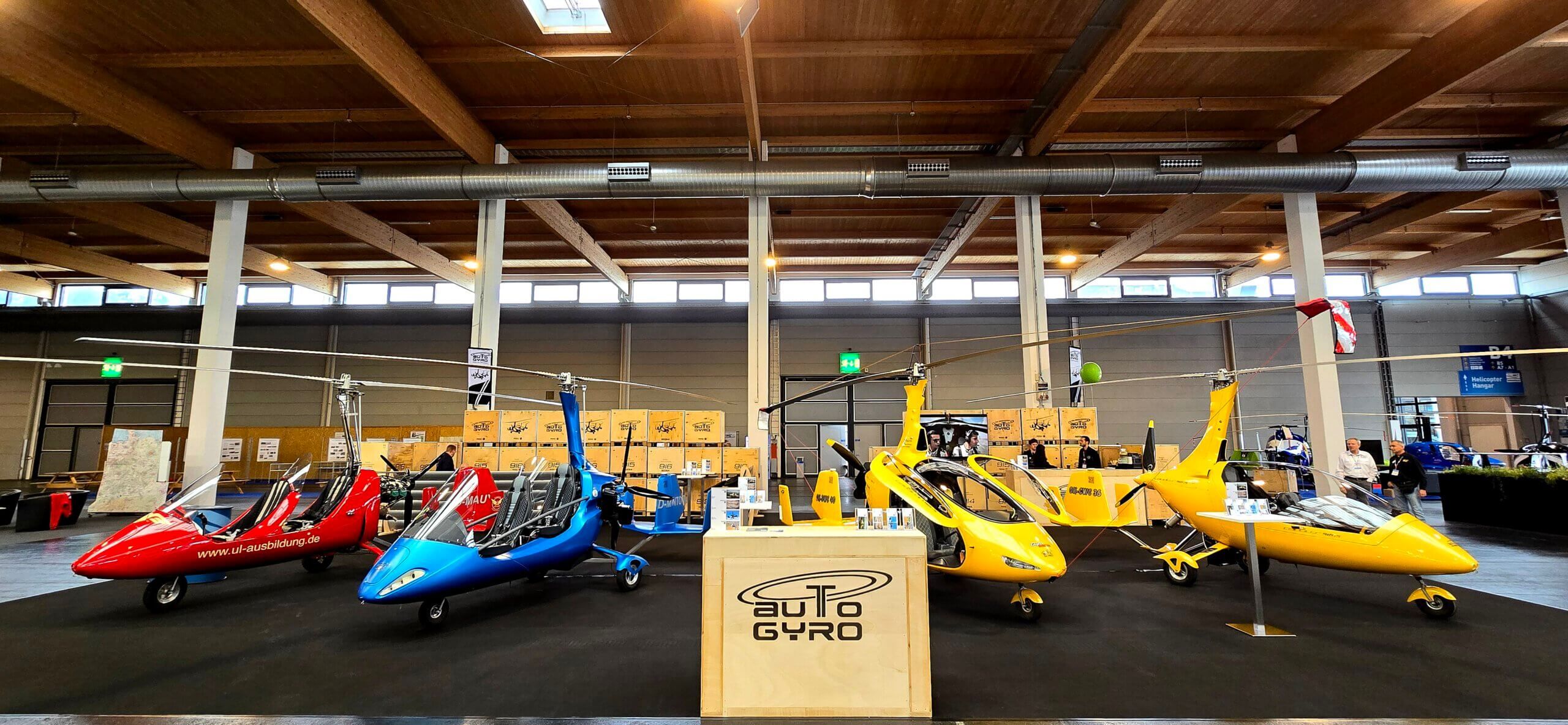 AutoGyro booth at the AERO 2024. In the front is a front desc with an AutoGyro logo and four gyroplanes in red, blue and yellow colour.