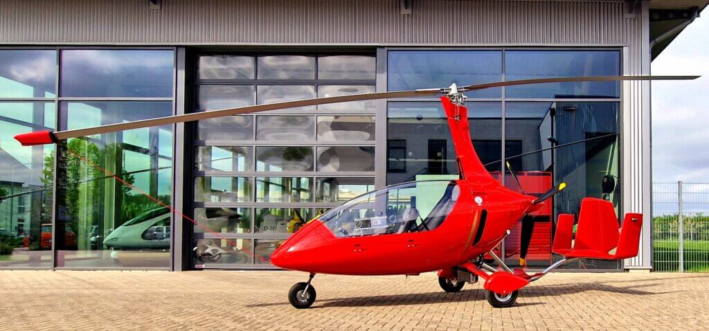 AutoGyro A red gyrocopter is parked on a paved area in front of a modern building with large reflective windows, marking the successful certifications achievement